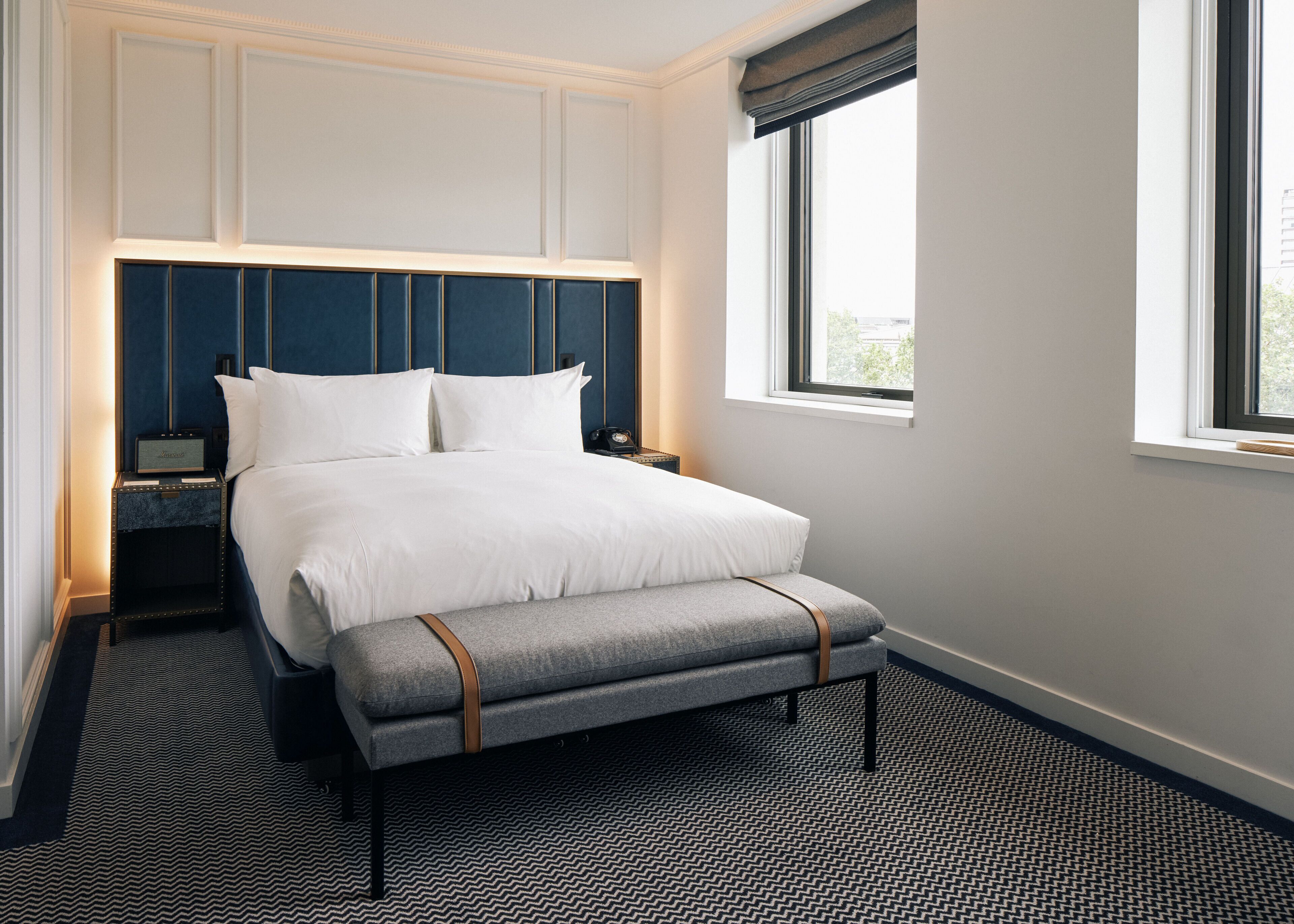 Una cama bien hecha con sábanas blancas en una habitación de hotel con cabecera azul oscuro, una mesita de noche pequeña y una ventana con vistas a la vegetación.
