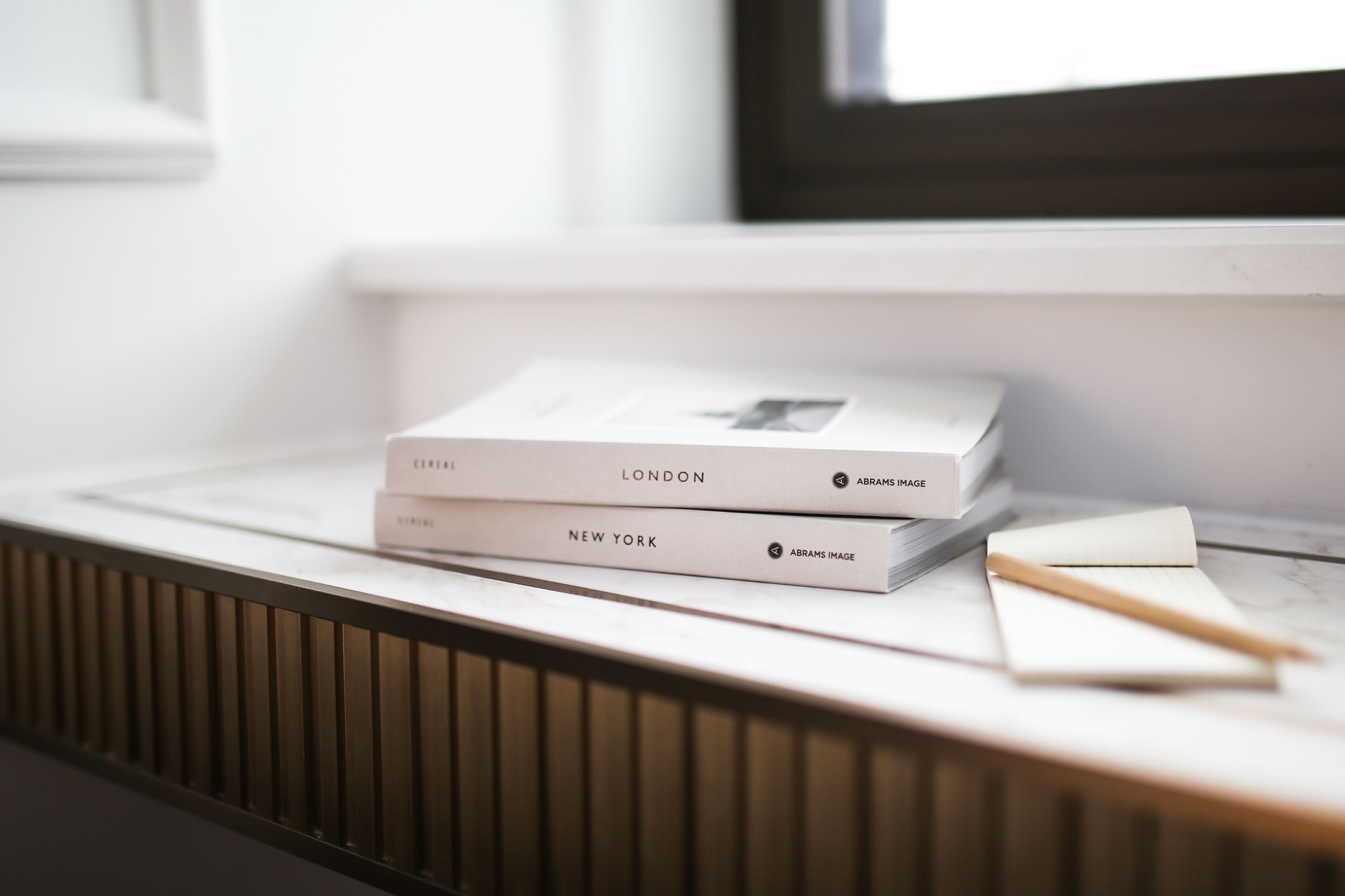 A stack of books titled 'Cereal', 'London', and 'New York' on a white surface near a window.