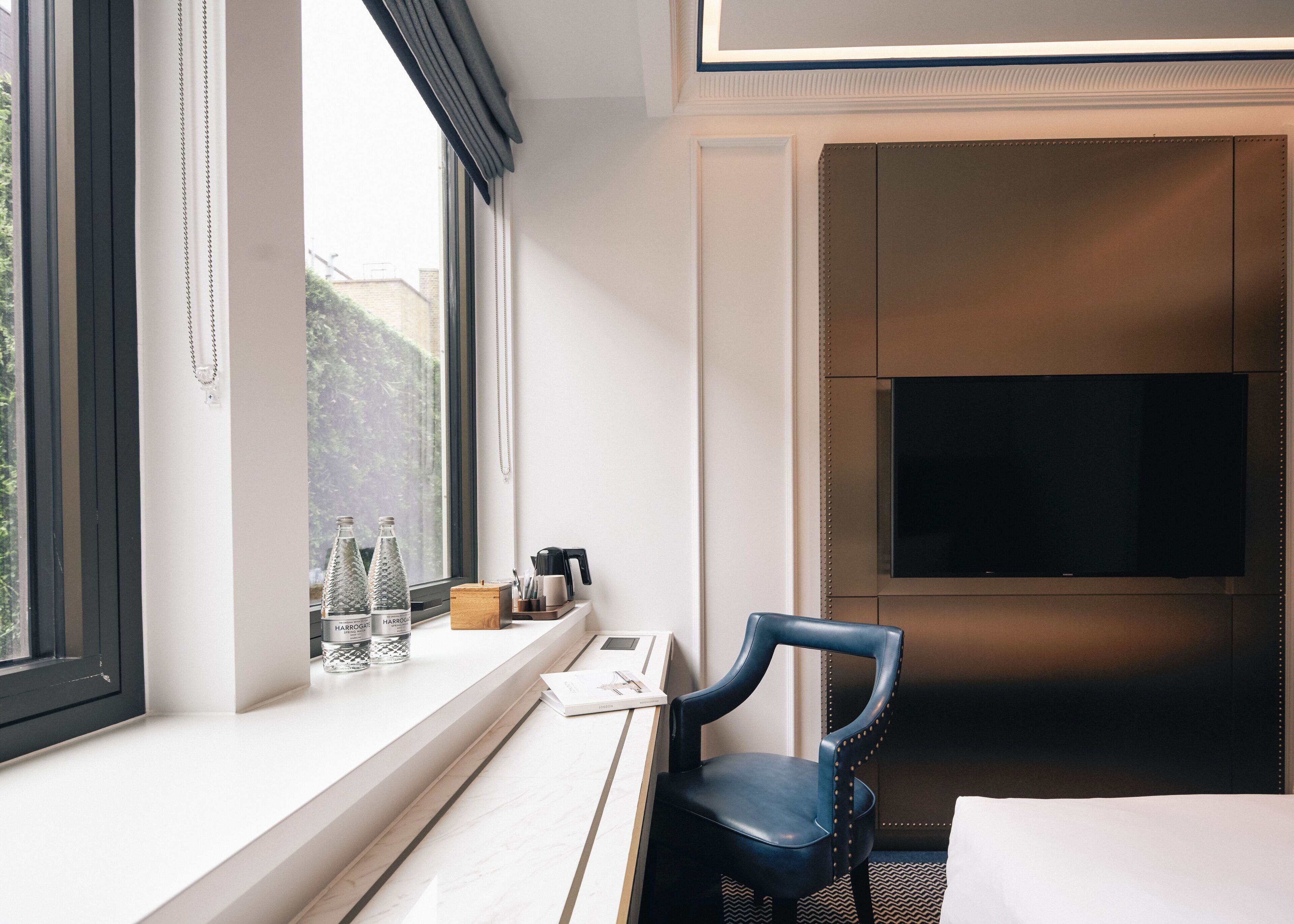 A hotel room with a large window, a flat-screen TV mounted on the wall, a desk with a chair, and a bed.