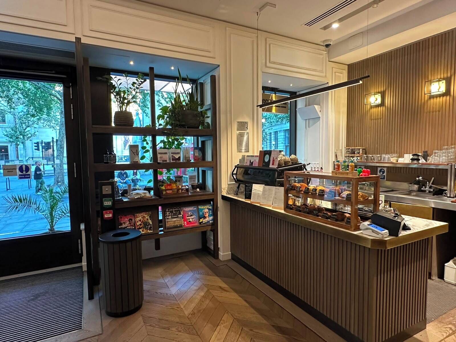 A modern cafe interior with a wooden counter, shelves with books and plants, and a view of the street outside.