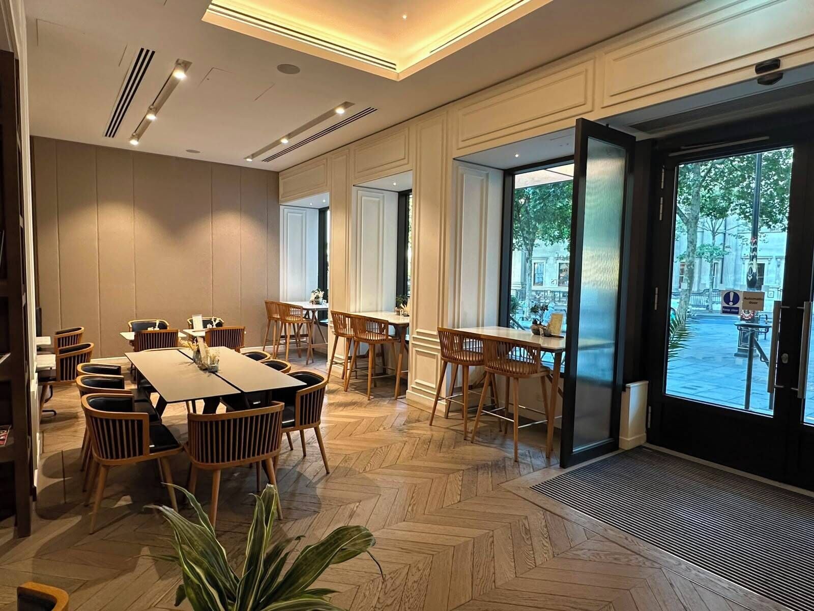Un comedor de hotel con varias mesas y sillas, ventanas amplias y un suelo con estampado de espiga.