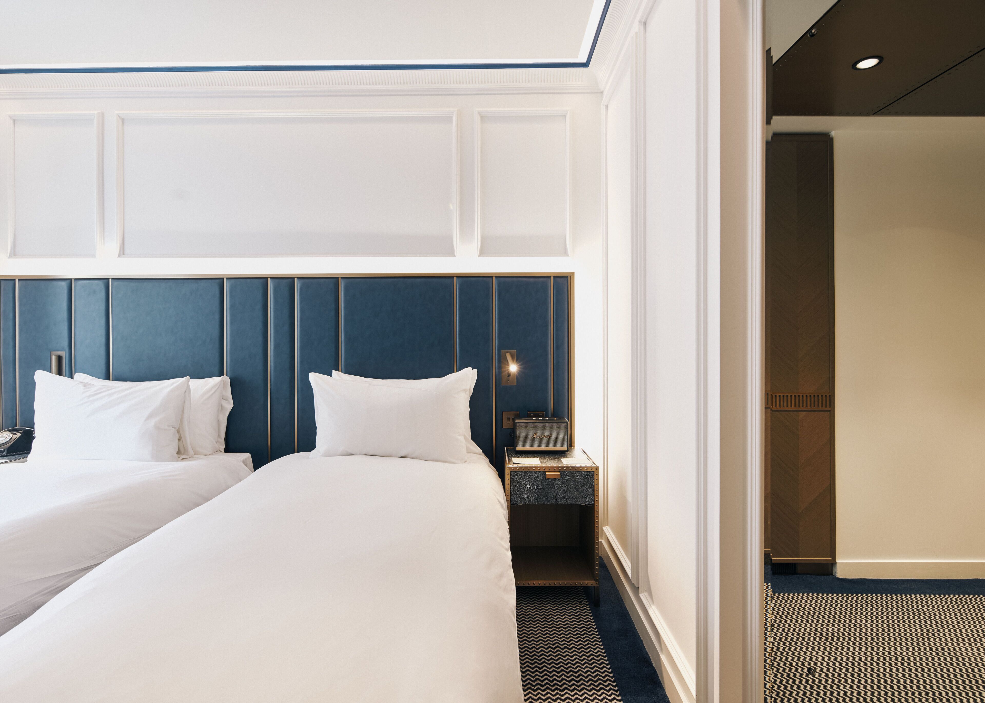 Habitación de hotel con dos camas, una mesita de noche con lámpara y vista al pasillo.
