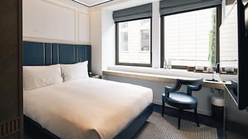Quarto de hotel com uma cama grande, uma mesa com cadeira, uma janela com vista para edifícios e um radiador sob a janela.