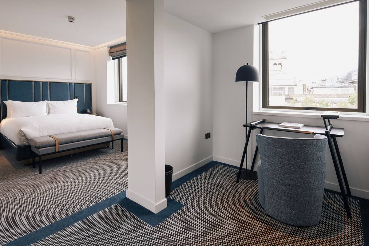 A hotel room with a bed, a desk, a lamp, and a view of the city through a large window.