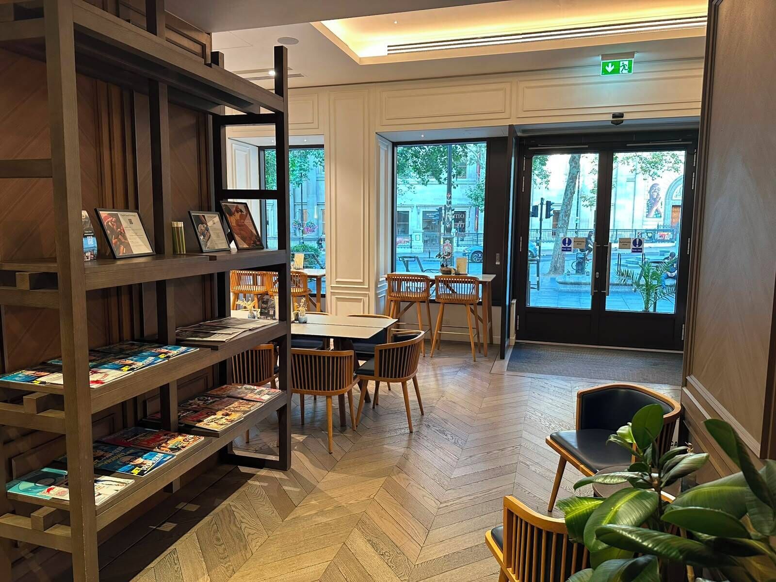 A library with bookshelves, tables, chairs, and a large window with a view of the street.