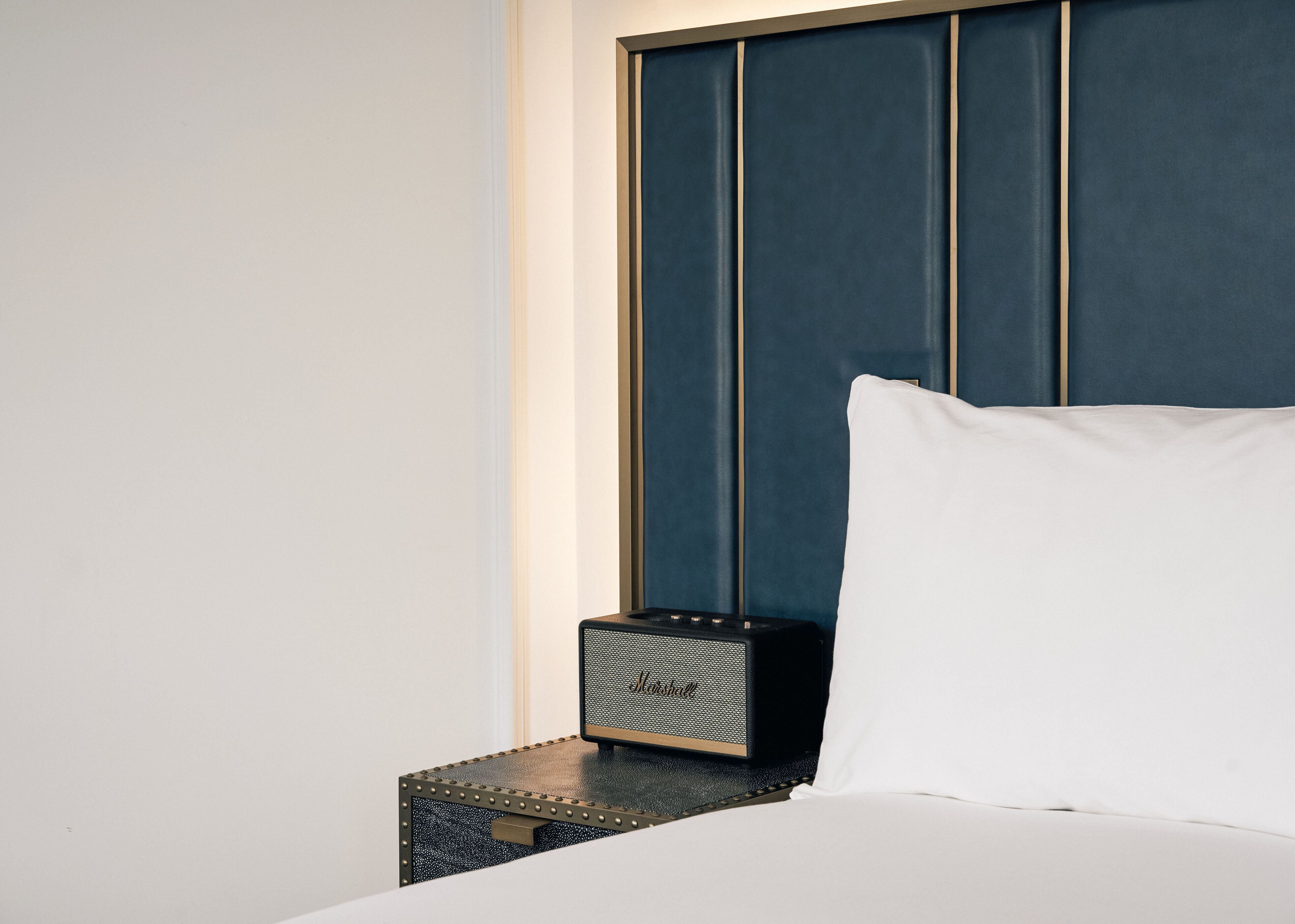 A neatly made bed with a white pillow and a black speaker on a textured side table.