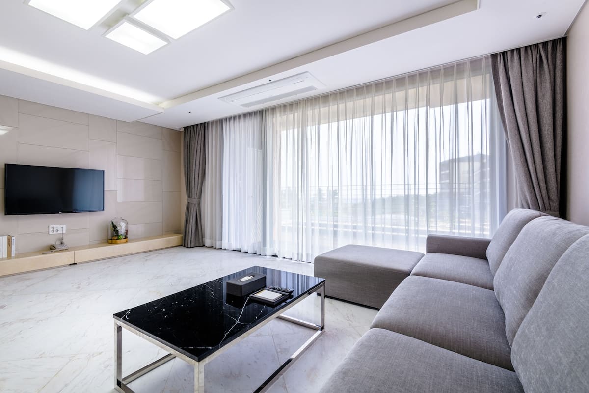 Ein modernes Wohnzimmer mit Flachbildfernseher, einem Couchtisch aus schwarzem Marmor, einem grauen Sofa und durchsichtigen Vorhängen am großen Fenster.