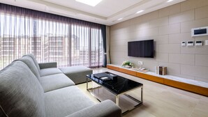 A modern living room with a sectional sofa, a black coffee table, a flat-screen TV mounted on the wall, and a window with sheer curtains. - Seaside Arden (Seogwipo)