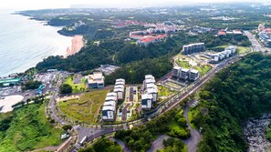 An aerial view of a coastal area with a beach, a parking lot, and several buildings with modern architecture. - Seaside Arden (Seogwipo)