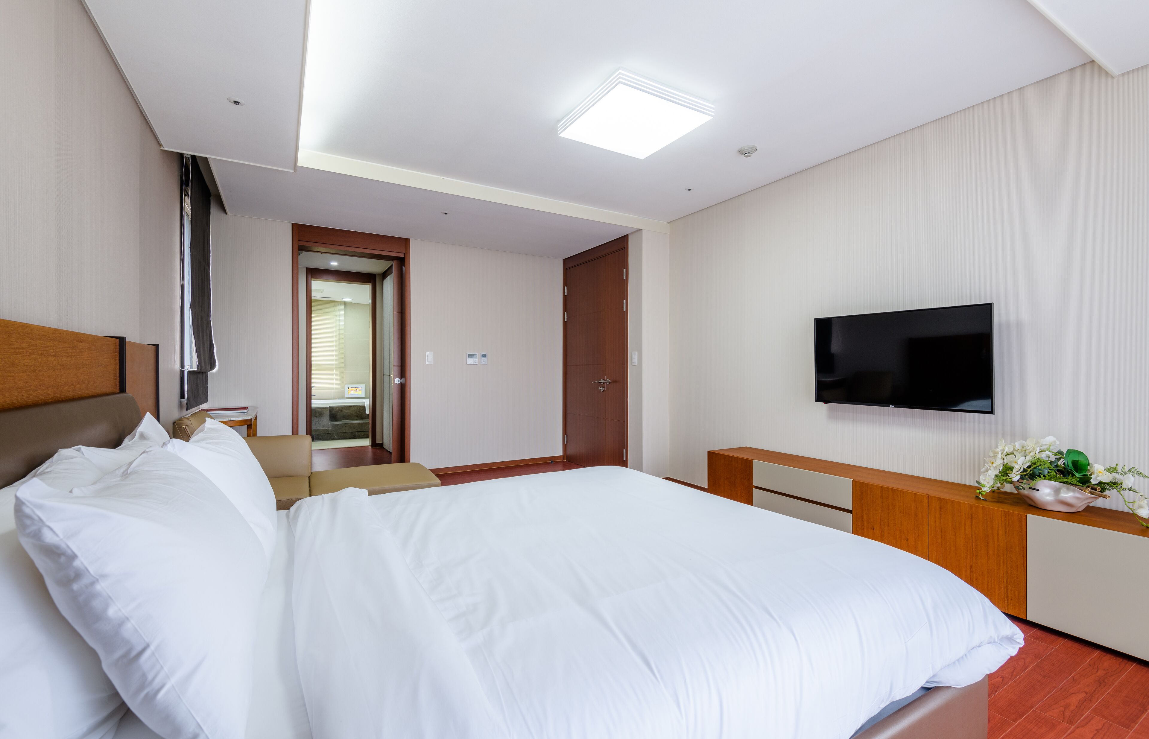 Habitación de hotel con una cama grande, un televisor de pantalla plana montado en la pared, un cabecero de madera, una mesita de noche y una pequeña planta en el tocador.