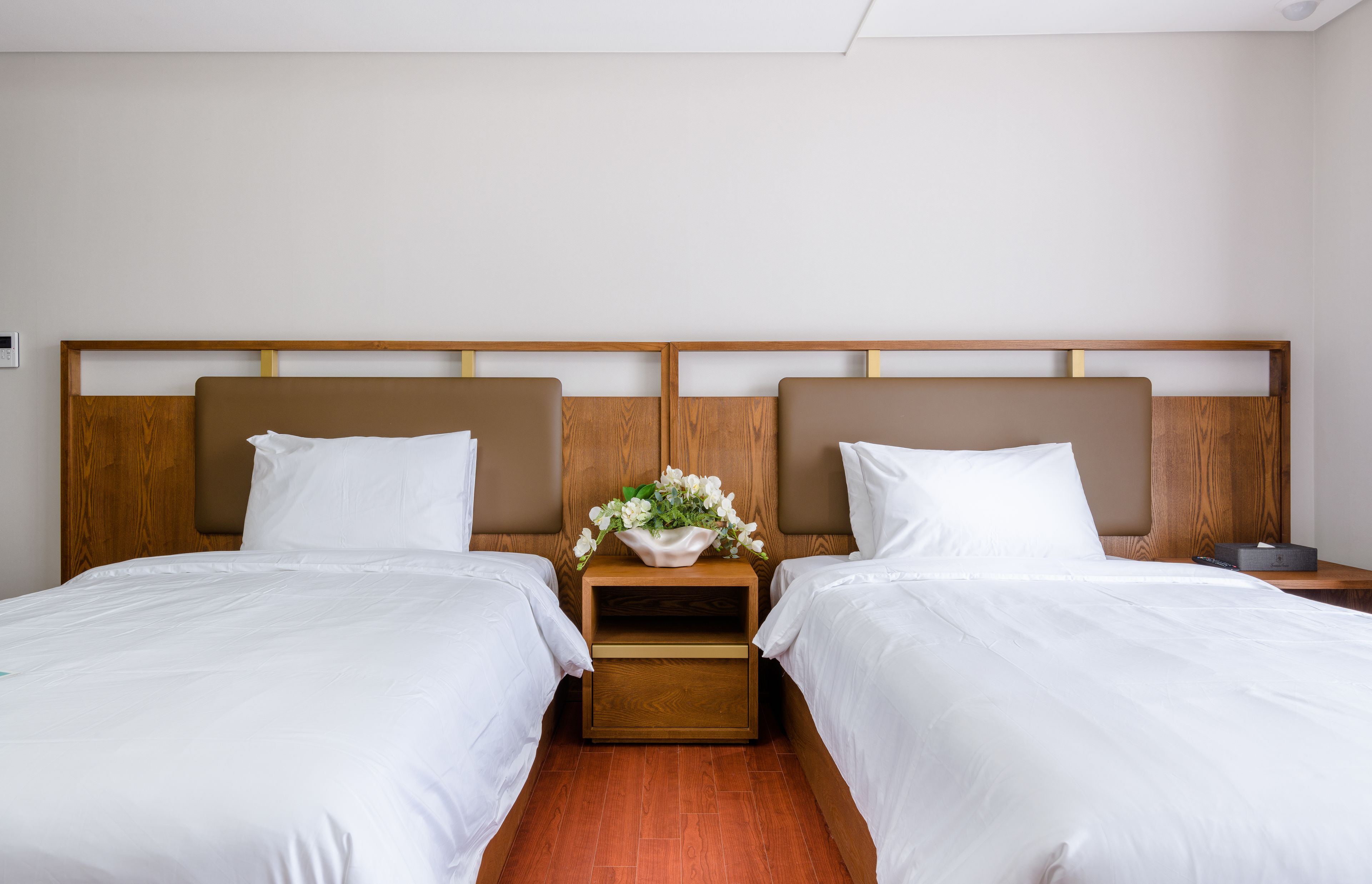 Habitación de hotel con dos camas, cabeceros de madera, ropa de cama blanca y una mesita de noche con un arreglo floral.