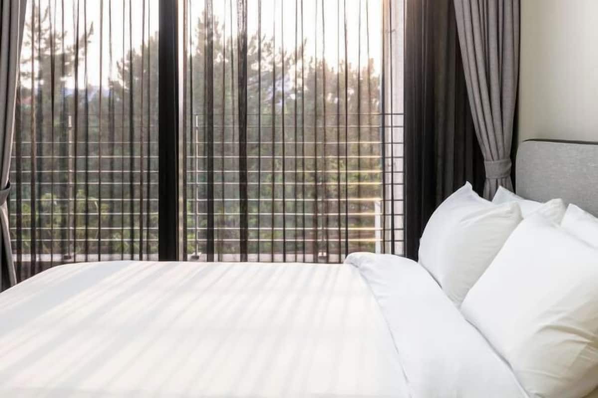 Ein Bett mit weißer Bettwäsche, einem grauen Kopfteil und Blick durch vertikale Jalousien auf ein bewaldetes Gebiet.