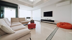 Een moderne woonkamer met een bank, poef en een traagschuimkussen, een televisie en een groot raam dat voor daglicht zorgt.