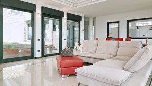 Een ruime woonkamer met een grote beige bank, een rode poef en een Louis Vuitton tas op de poef. Er zijn grote ramen met uitzicht op een gebouw.