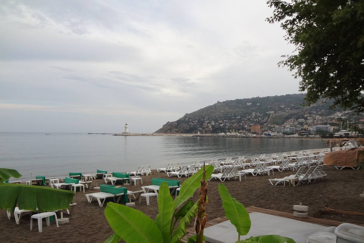 Une zone de plage aménagée avec des chaises longues et des tables, un phare visible au loin et une ville perchée sur une colline en arrière-plan.