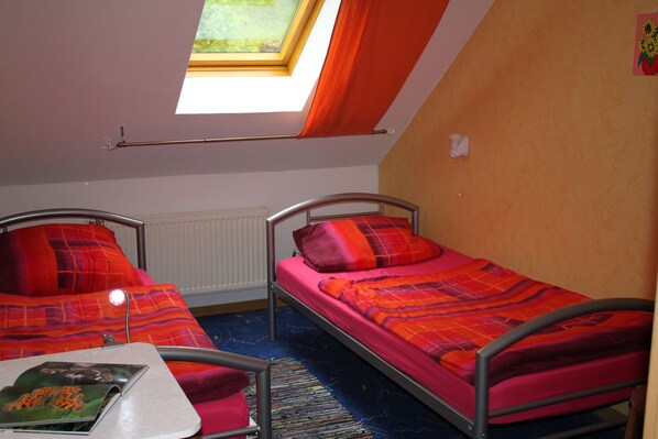 A room with two metal bunk beds, each with a red and orange blanket. There is a skylight above the beds, a radiator on the wall, and a small table with a book. - Ferienwohnungen Hees (Emmerzhausen)
