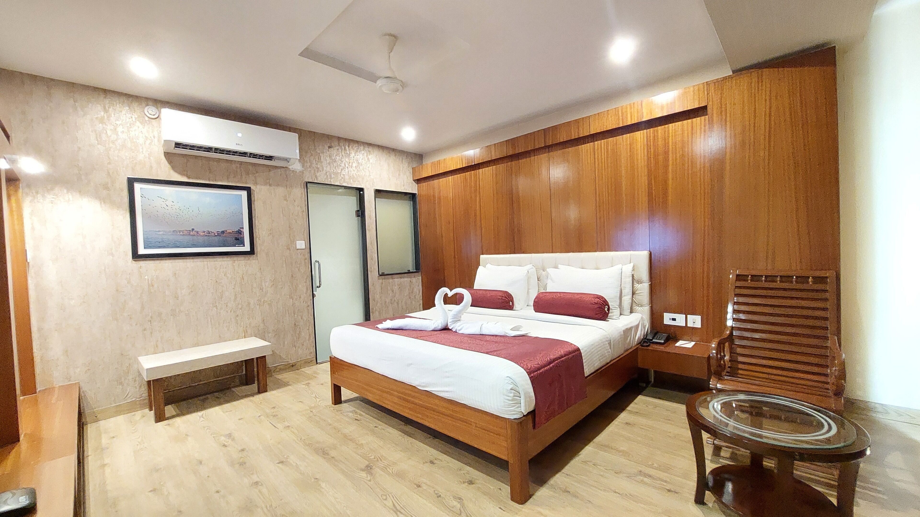 A modern hotel room with a wooden bed, a bedside table, a small round table, and a large window.