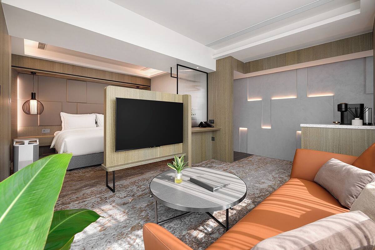 A modern hotel room with a large bed, a flat-screen TV, a round coffee table, and a seating area with orange cushions.