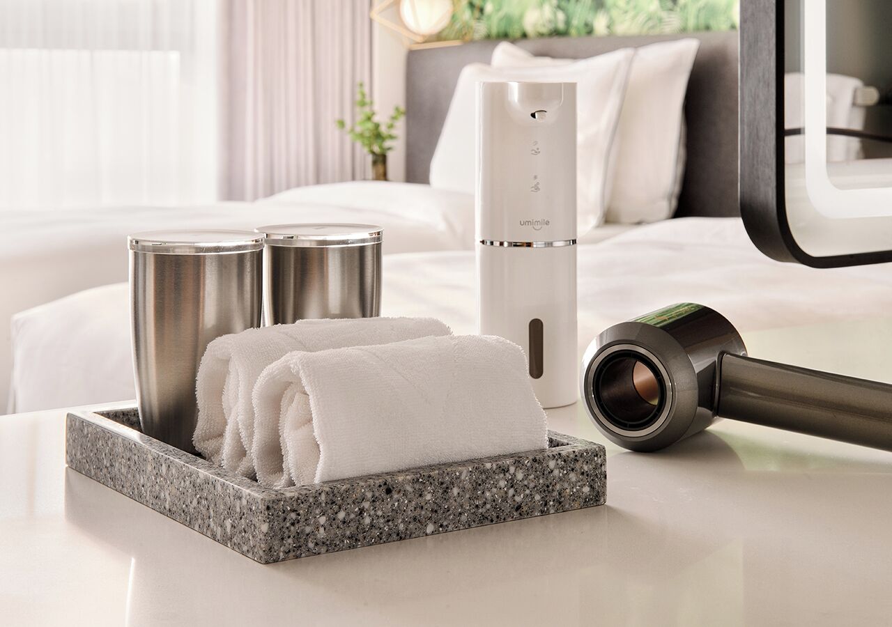 Une chambre d’hôtel moderne avec une salle de bain bien aménagée, comprenant un lavabo en granit, un porte-serviettes avec des serviettes pliées, une douchette à main blanche et une douche électrique blanche compacte.