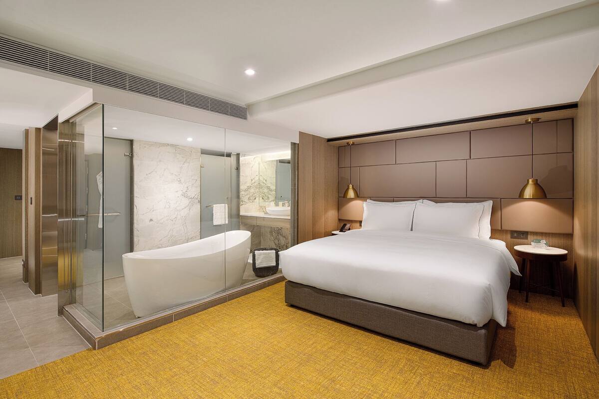 A modern hotel room with a large bed, a glass-enclosed shower, a bathtub, and a small bedside table with a lamp.