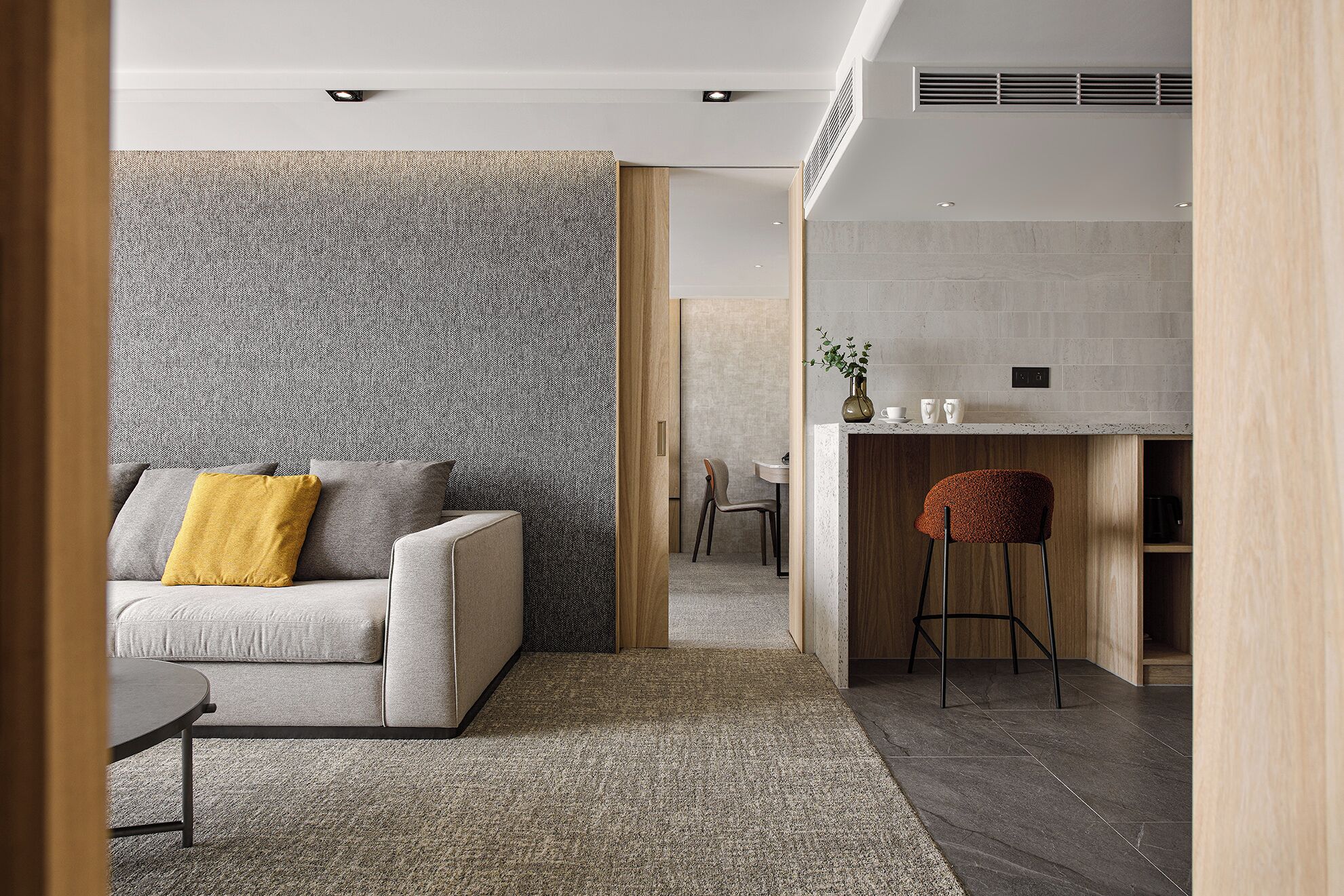 A modern living room with a grey sofa, a patterned wall, and a kitchen area with a bar stool.
