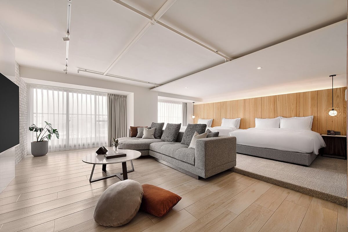 A modern hotel room with a large bed, a grey sofa, a round coffee table, and a potted plant.