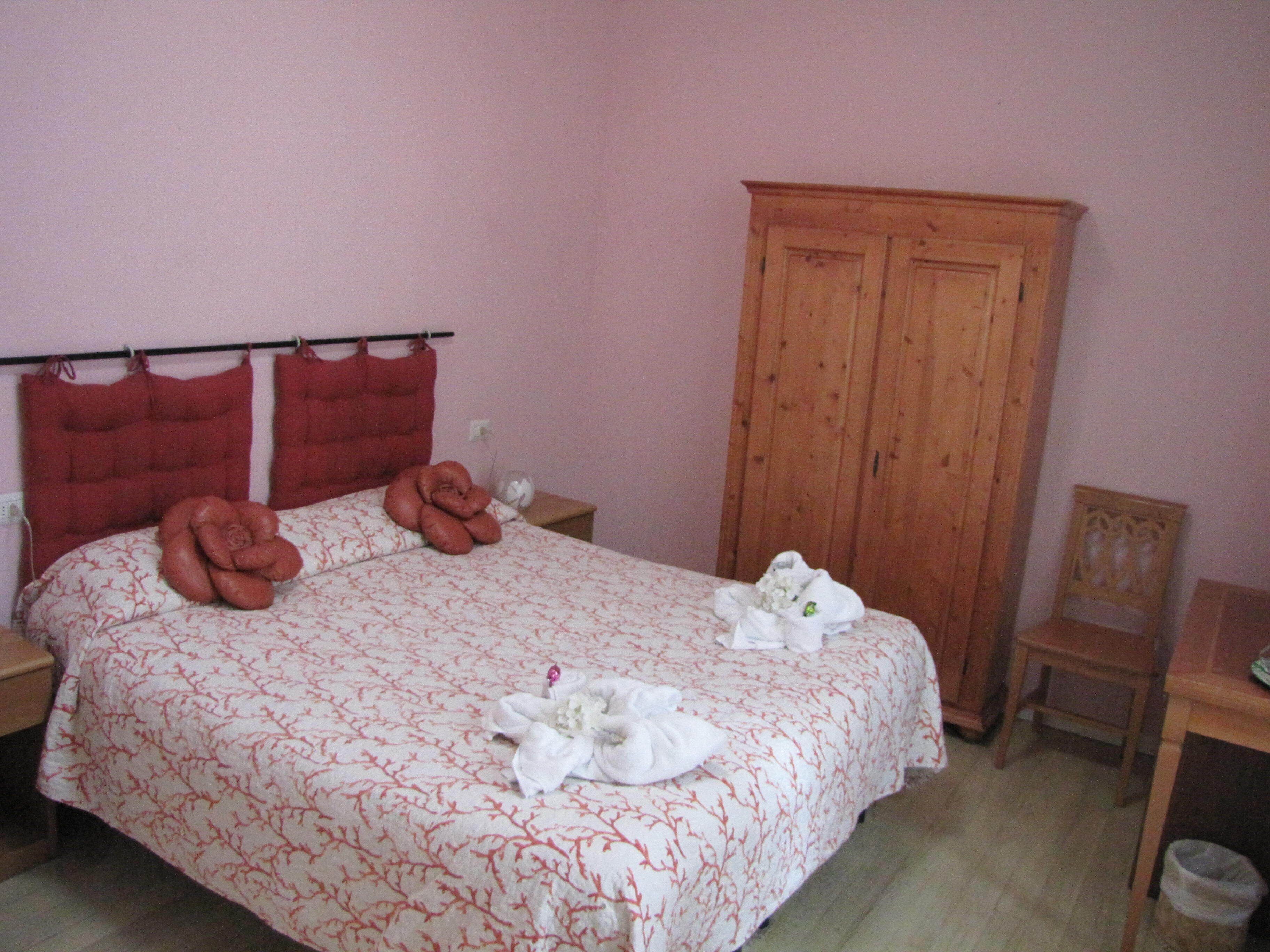 A neatly made bed with towels and a red headboard. There is a wooden wardrobe and a small table with a chair.