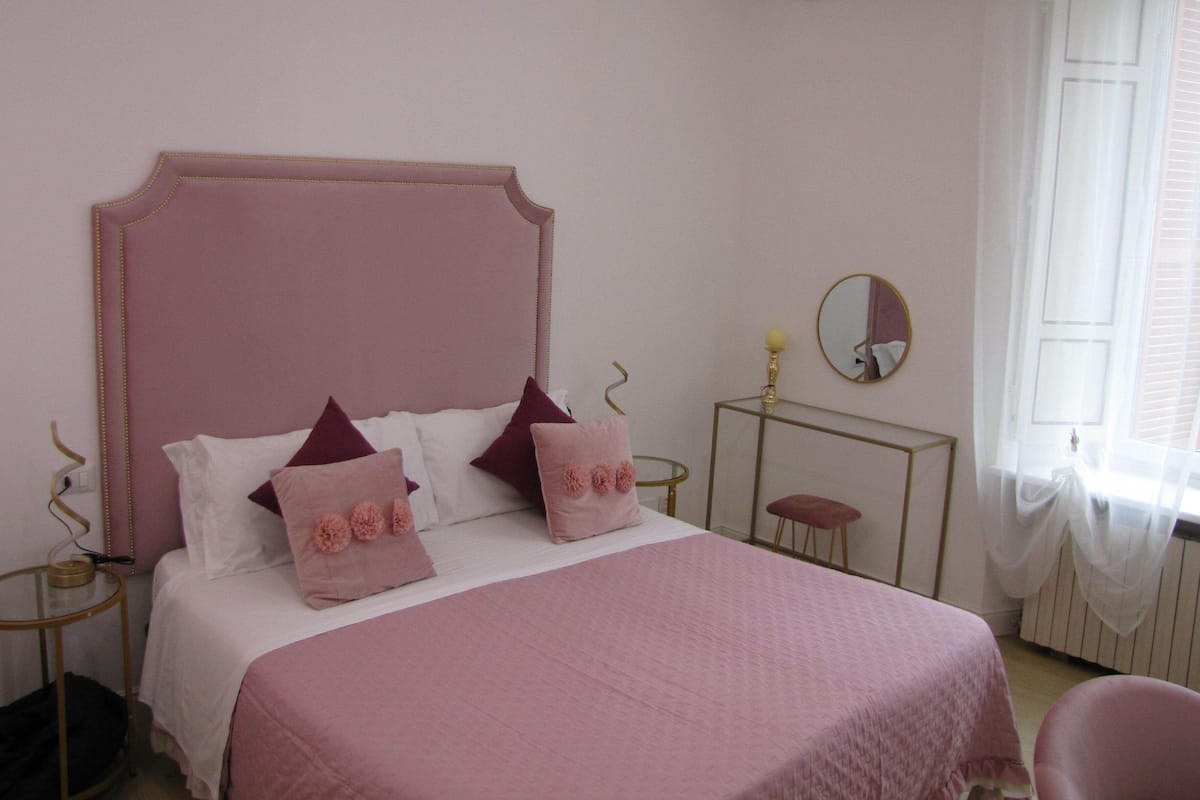 A bedroom with a pink bedspread, white pillows, and a mirror on a bedside table.