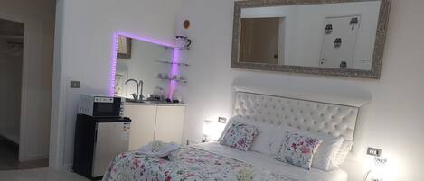 A bedroom with a bed, a bedside table, a mirror, and a kitchenette area.