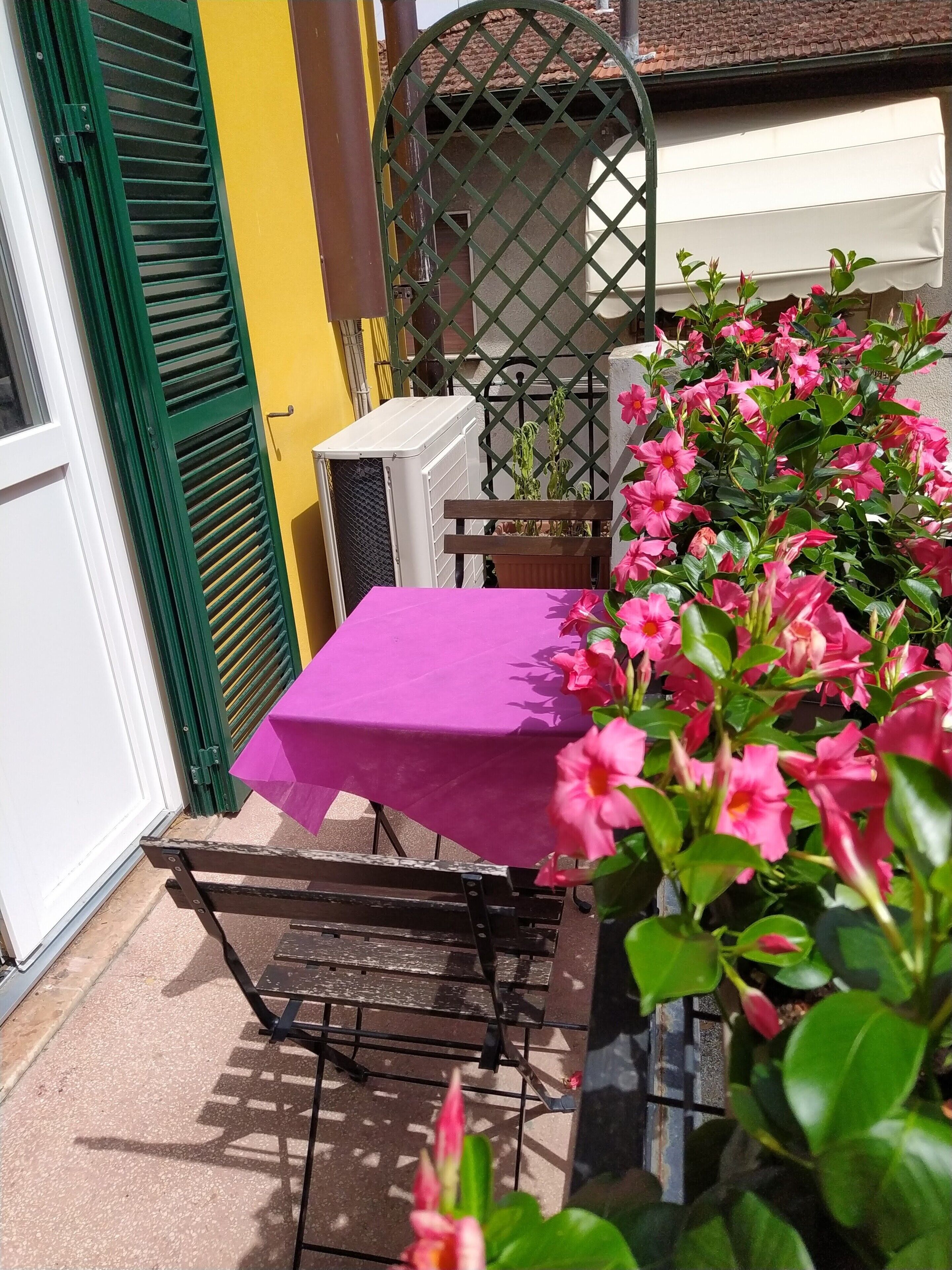 Patio esterno con tavolo ricoperto da una tovaglia viola, una sedia in metallo e un vaso con fiori rosa.