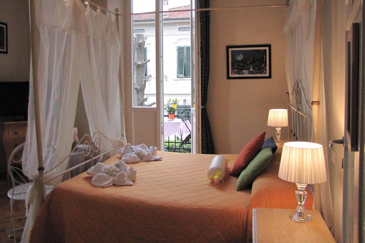 A hotel room with a bed, a bedside table with a lamp, a window with a view of the outside, and a door.