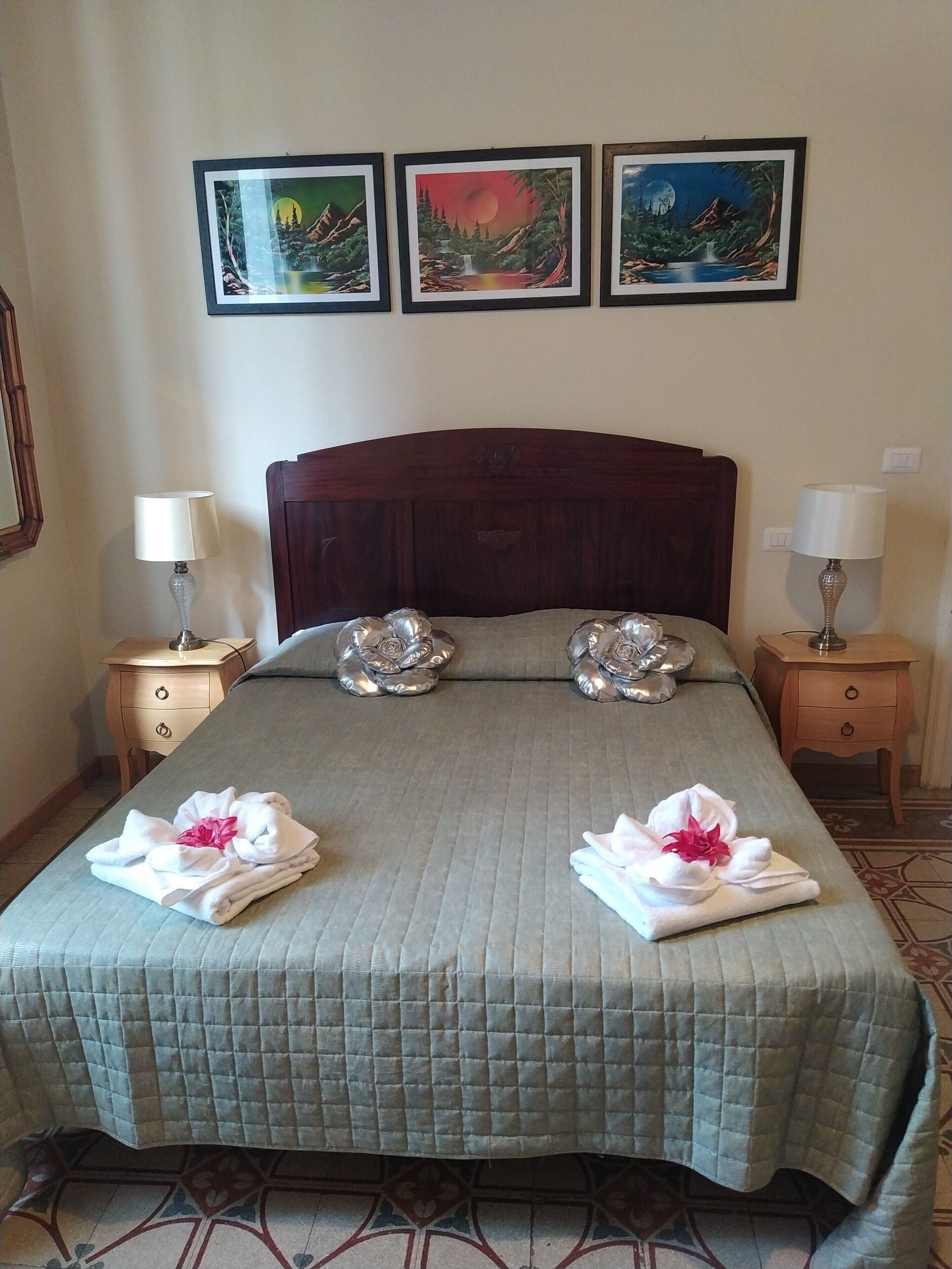 A neatly made bed with a quilted cover, two decorative pillows, and two towels with flowers on each side. There are three framed pictures on the wall above the bed.