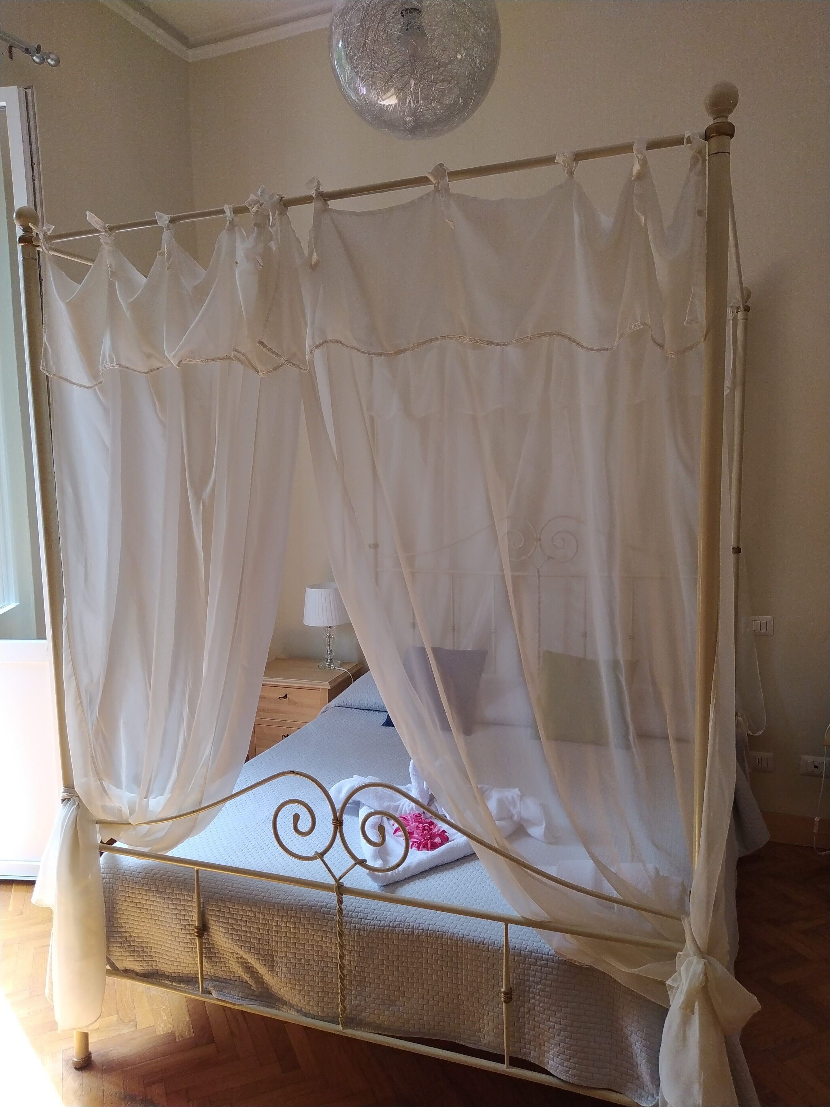 Uma cama com dossel e cortinas leves, uma mesa de cabeceira com abajur, e piso de madeira.