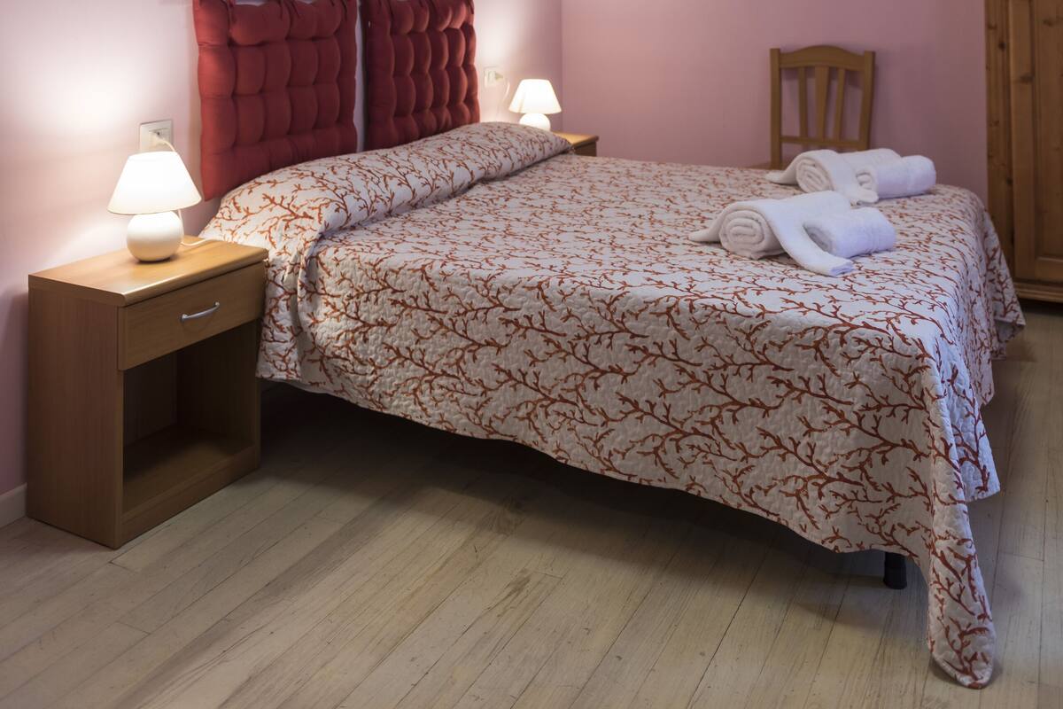 A bedroom with a bed, two bedside tables with lamps, a wooden chair, a wardrobe, and two paintings on the wall.