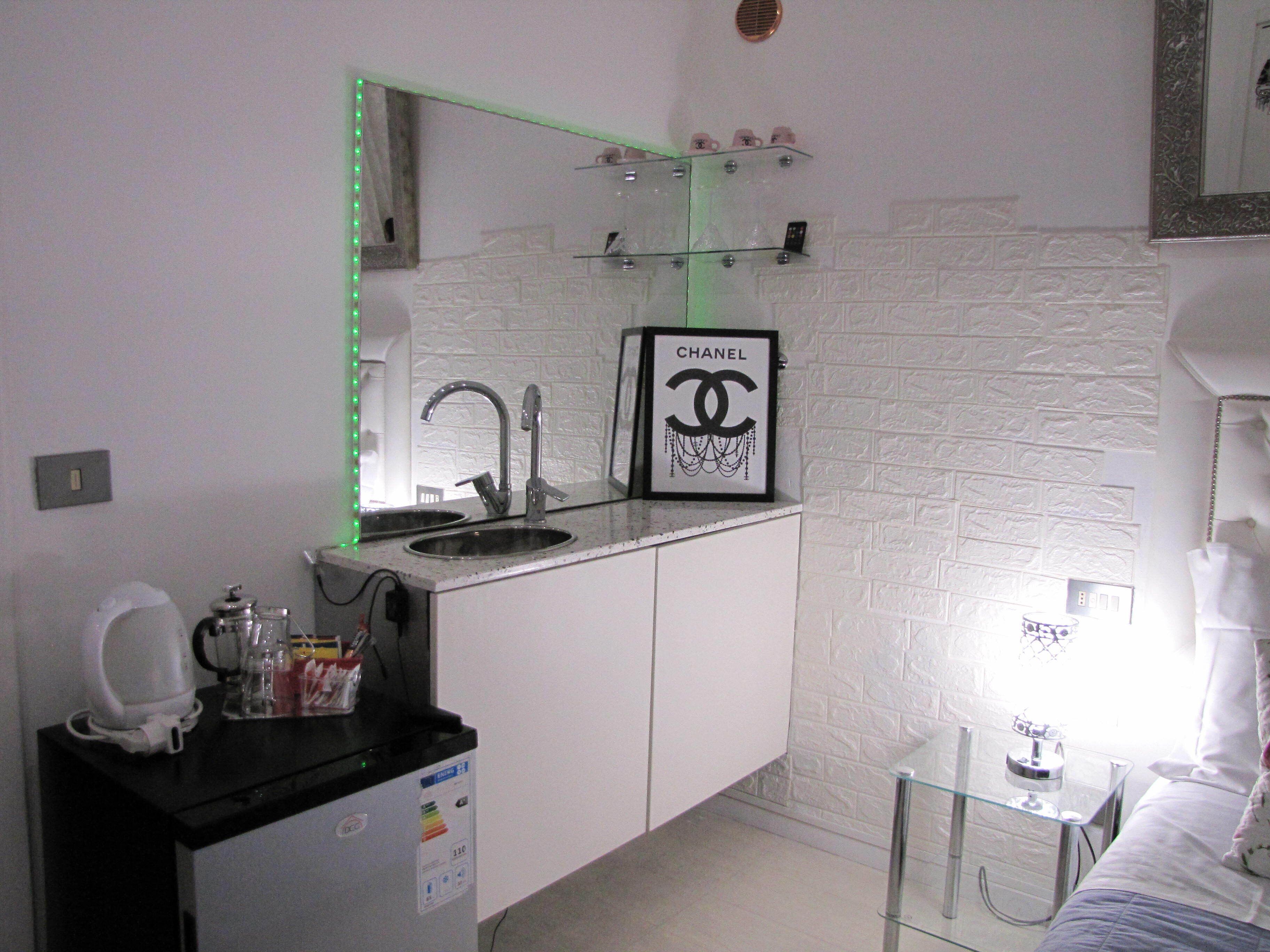 Un baño pequeño y moderno con pared de azulejos blancos, un lavabo de vidrio con grifo cromado, un espejo con luces LED verdes, un logotipo de Chanel enmarcado y una mesita de noche con lámpara.