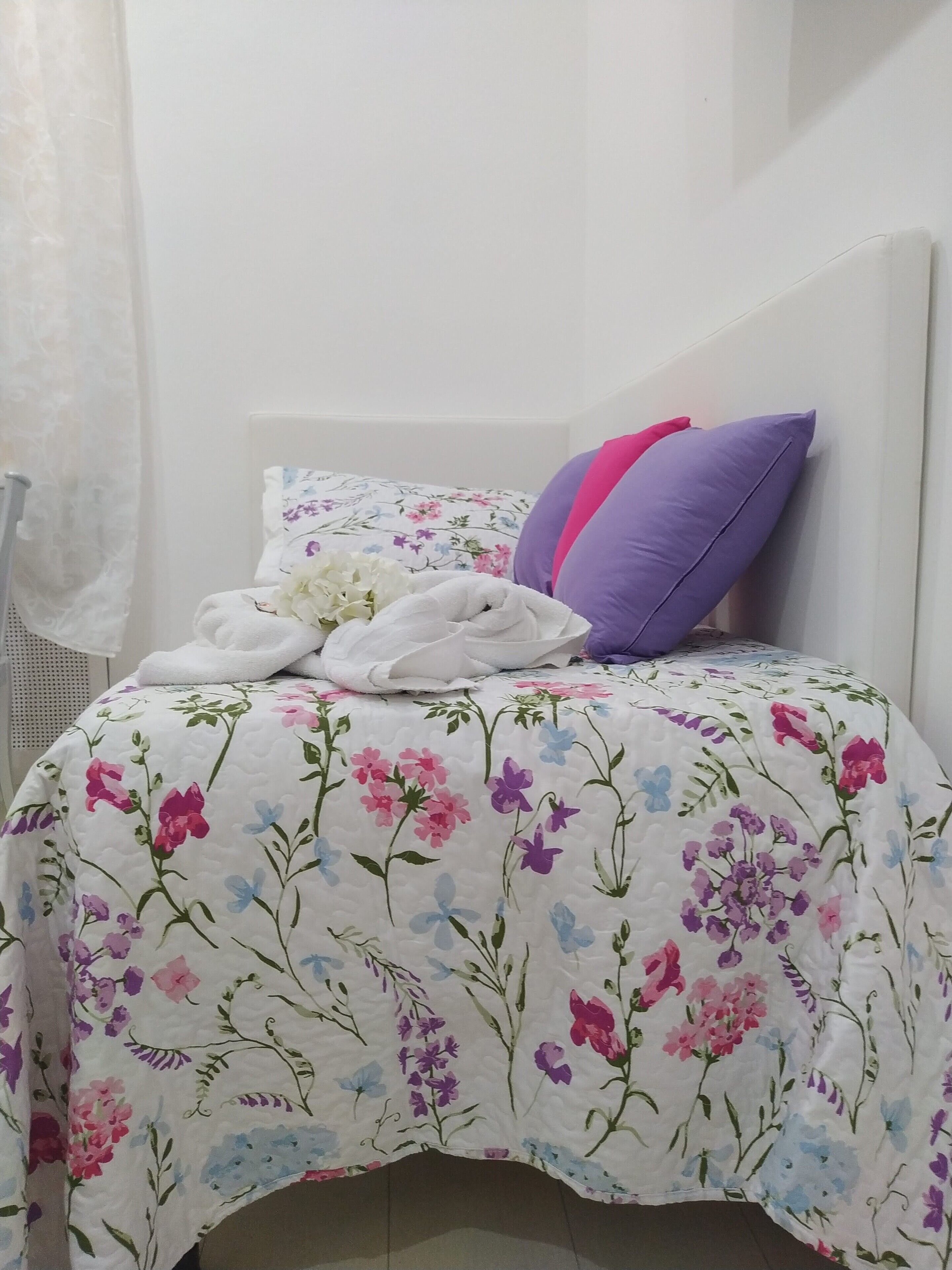A neatly made bed with a floral bedspread, a white towel, and two pillows, one purple and one pink.