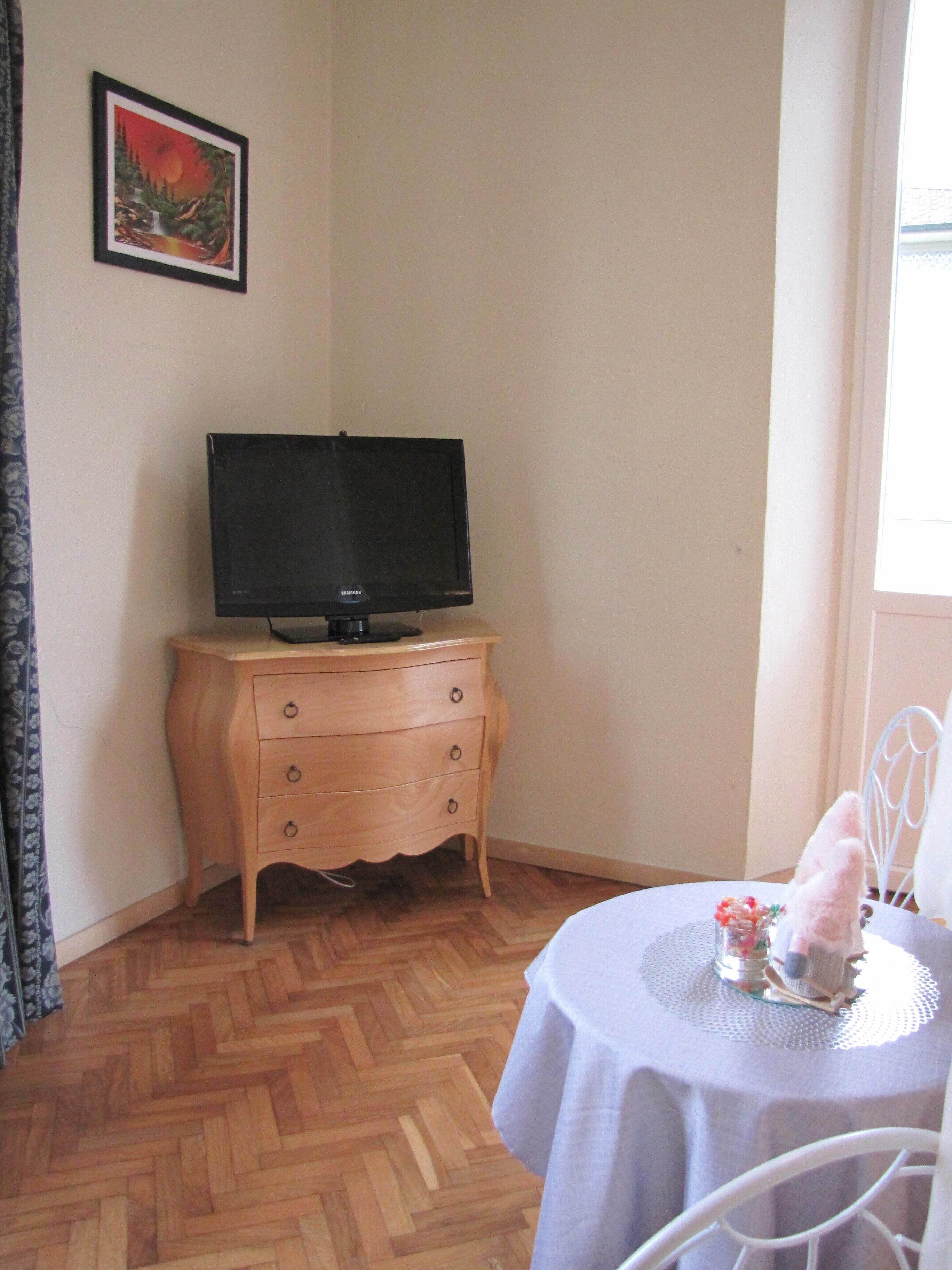 A room with a wooden dresser, a television on top, a small table with a decorative item, and a chair.
