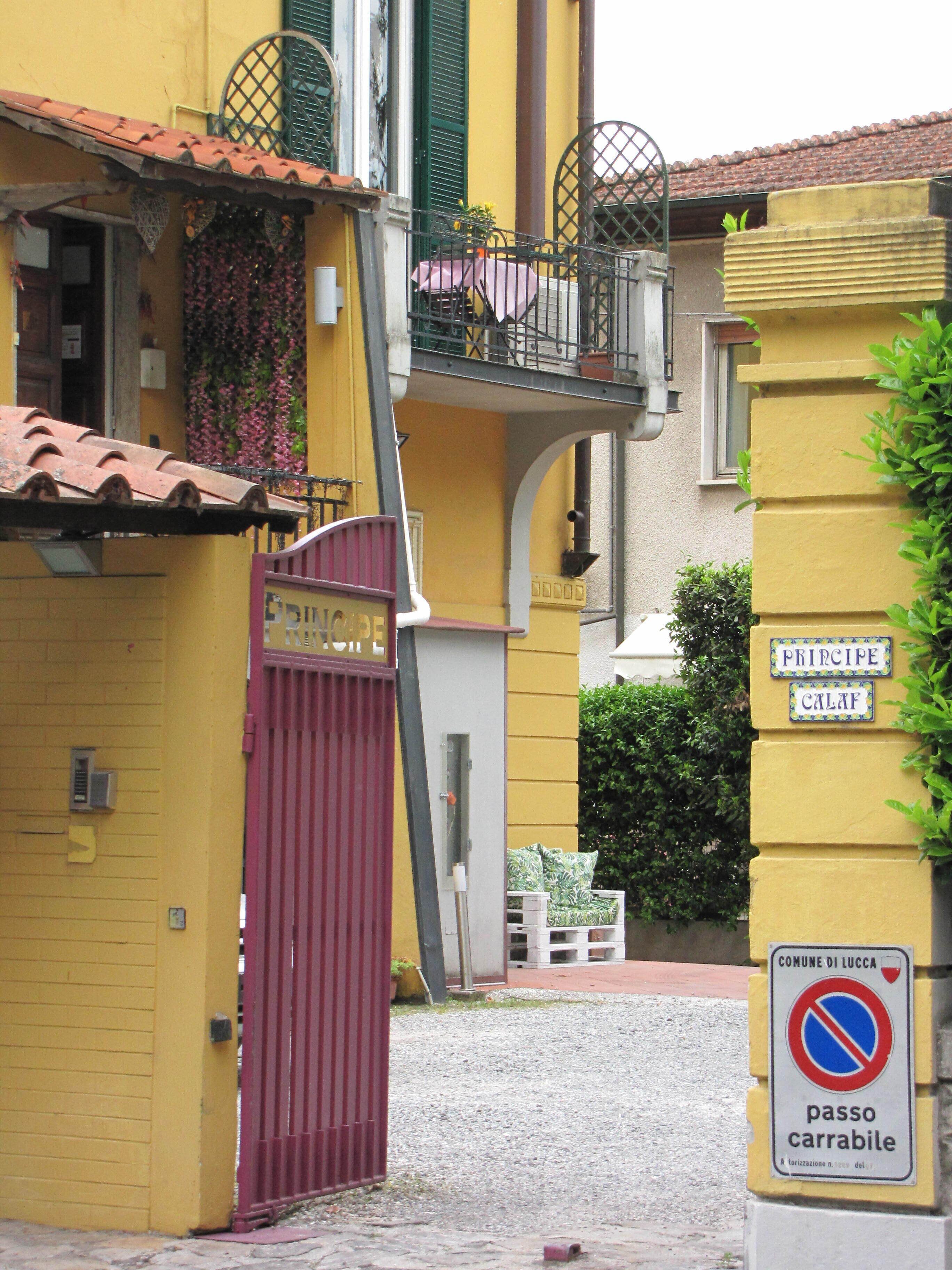 A yellow building with a red gate, a balcony with green shutters, and a sign indicating a no parking area.