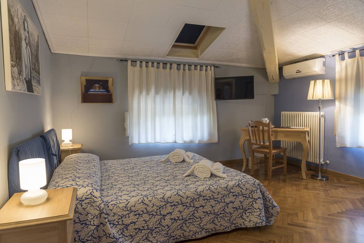A hotel room with a bed, a desk, a chair, a lamp, a radiator, and a window with curtains.
