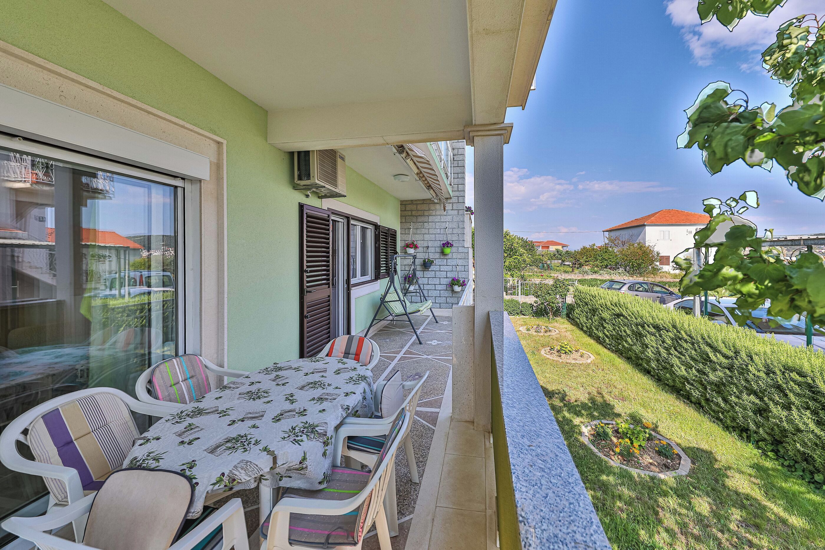 Ein Balkon mit Tisch und Stühlen, ein Fenster mit Jalousien und Blick auf einen Garten sowie parkende Autos.