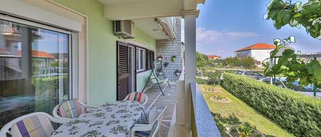Ein Balkon mit Tisch und Stühlen, ein Fenster mit Jalousien und Blick auf einen Garten sowie parkende Autos.