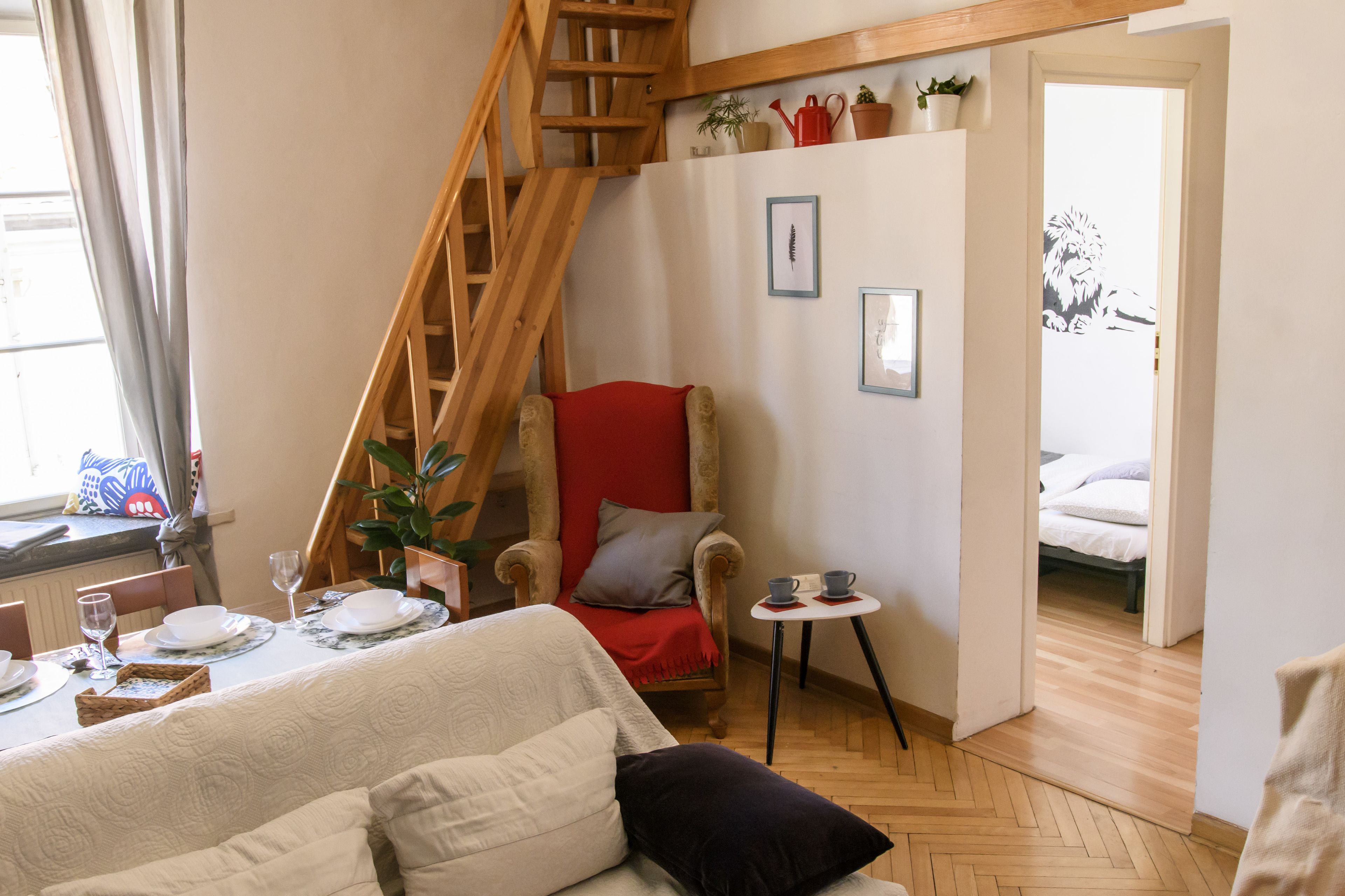 A cozy room with a wooden staircase, a red armchair, a small table with cups, and a bed in the adjacent room.