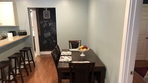 A dining area with a wooden table, four chairs, a fruit bowl, and a chalkboard with writing. - Nana's Cottage 3 miles from downtown, pet friendly (Asheville)