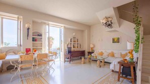 A spacious living room with a white sofa, a dining table with chairs, and a TV stand. There is a staircase with a chandelier and a plant hanging on the wall. - Solemar Sicilia - Casa Porto Bagnera (Santa Flavia)
