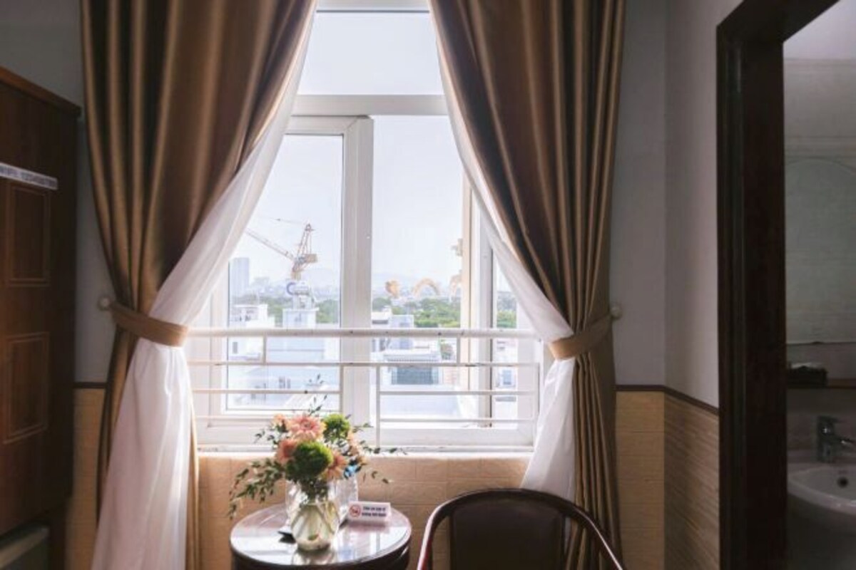 A room with a window, curtains, a small table with a vase of flowers, and a chair.