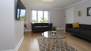Een moderne woonkamer met een bank, een glazen salontafel, een kroonluchter en een aan de muur bevestigde televisie.
