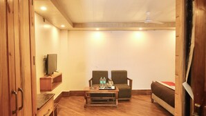 A room with wooden flooring, a sofa, a chair, a small table with bottles, and a television on a shelf. - Ashirwad Hotel and Spa (Mussoorie)