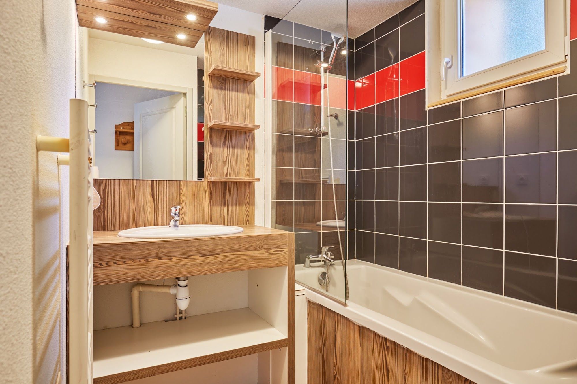 A modern bathroom with a wooden vanity, a large mirror, a white sink, a bathtub, and a shower enclosure with red and black tiles.