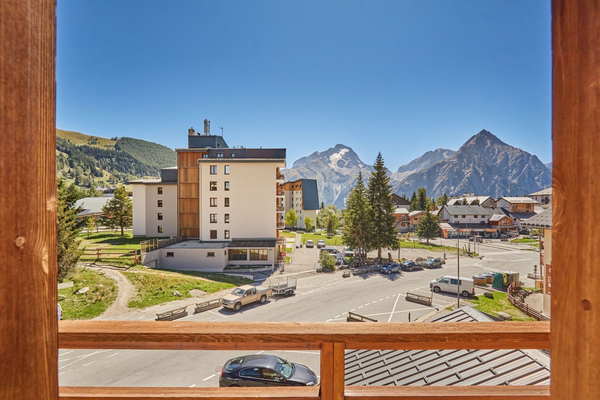 A view of a building with multiple floors and a parking area in front, with mountains in the background.