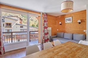 A room with a wooden interior, a gray sofa, a wooden table, and a view of buildings through large windows. - Remontées à 50 m | Piscine | Cuisine équipée (Les Deux Alpes)